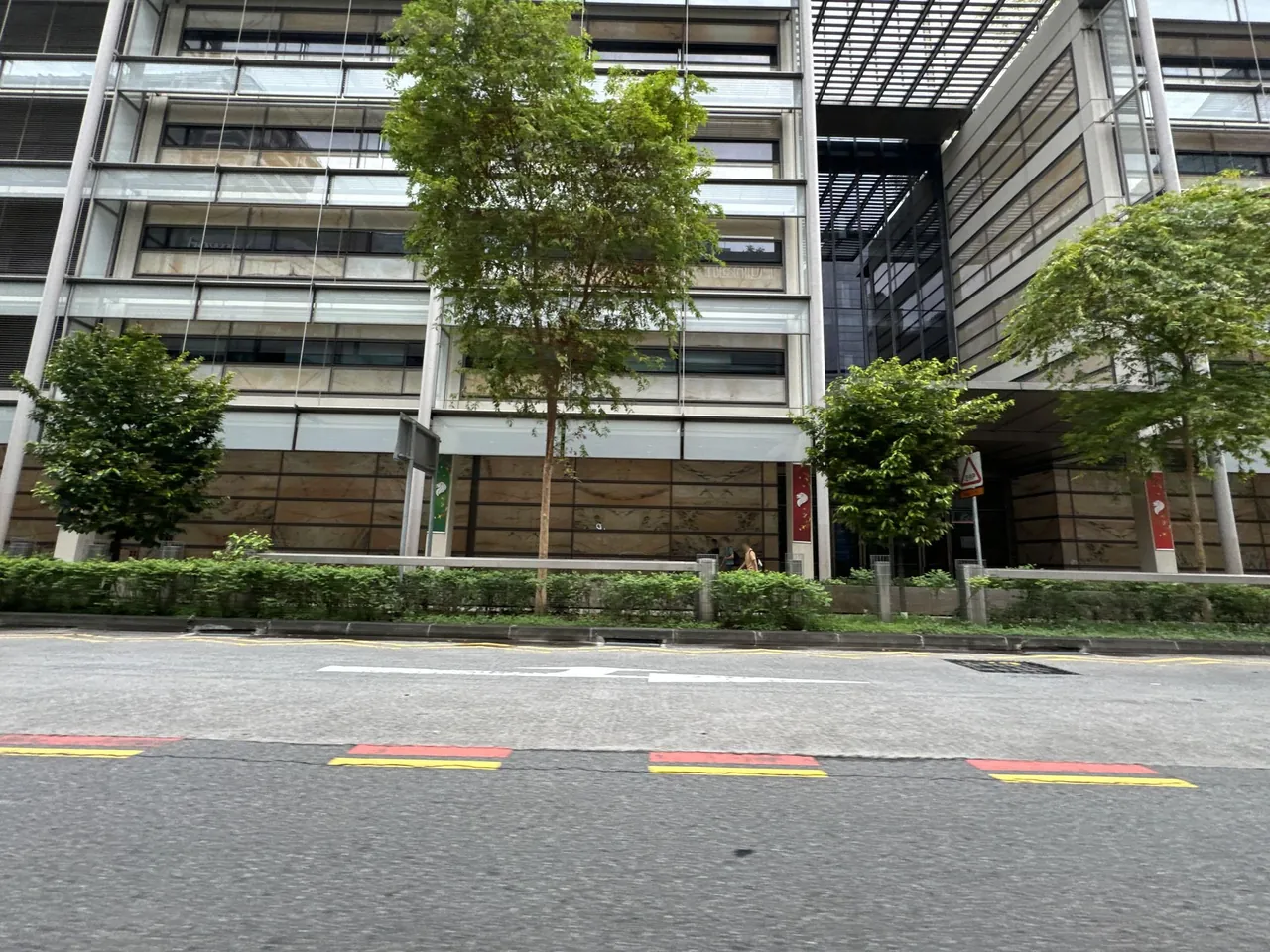 Short red-and-yellow road markings, a yellow edge line, street trees and modern multi-storey buildings.